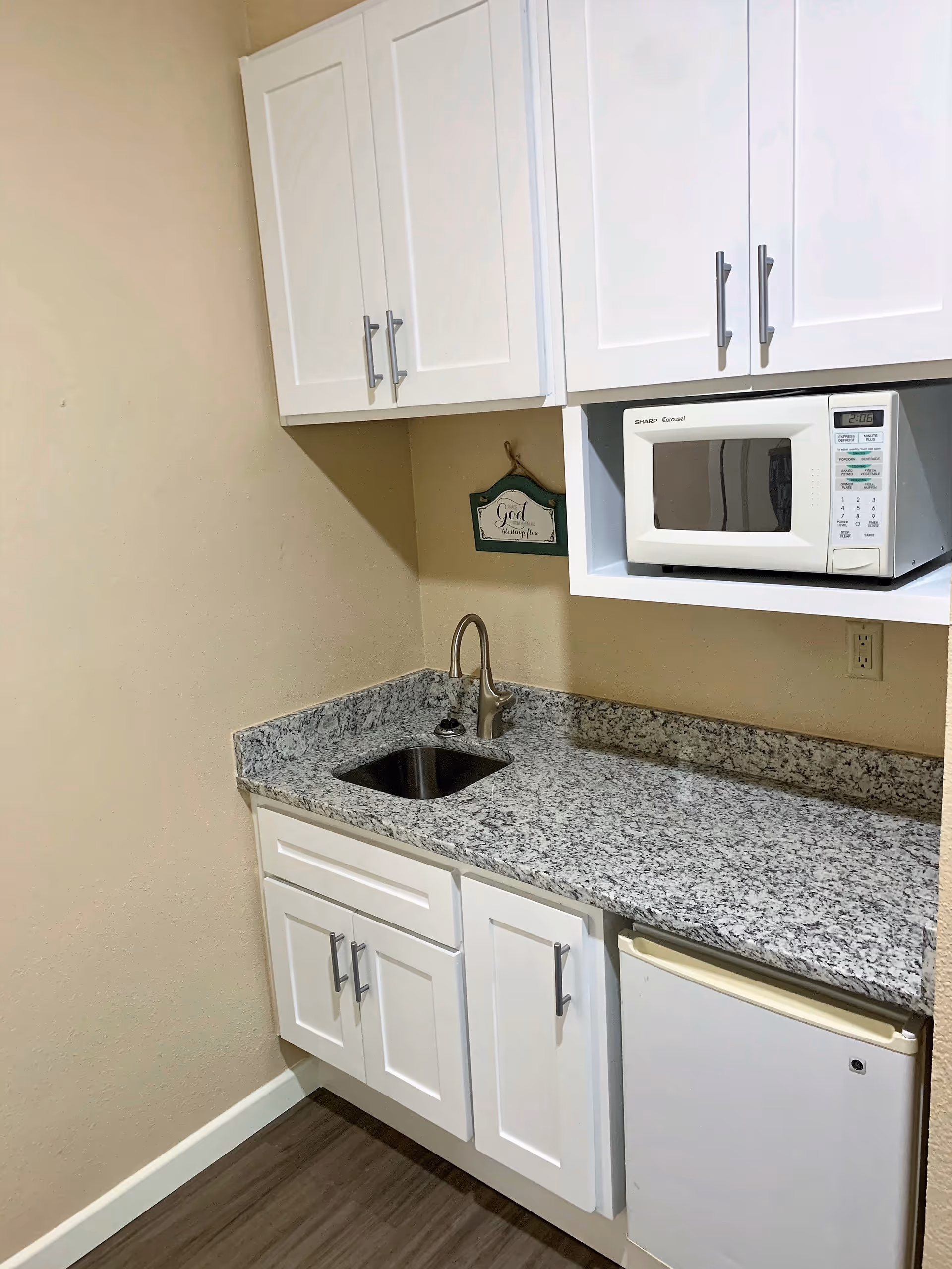Small kitchenette area with white cabinets, a granite countertop, a stainless steel sink with a faucet, a white microwave oven mounted above the counter, and a small white refrigerator below the counter. A decorative sign is hanging on the wall behind the sink.
