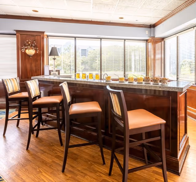 A bright communal bar area with a granite countertop, four high-backed stools, and large windows with blinds.