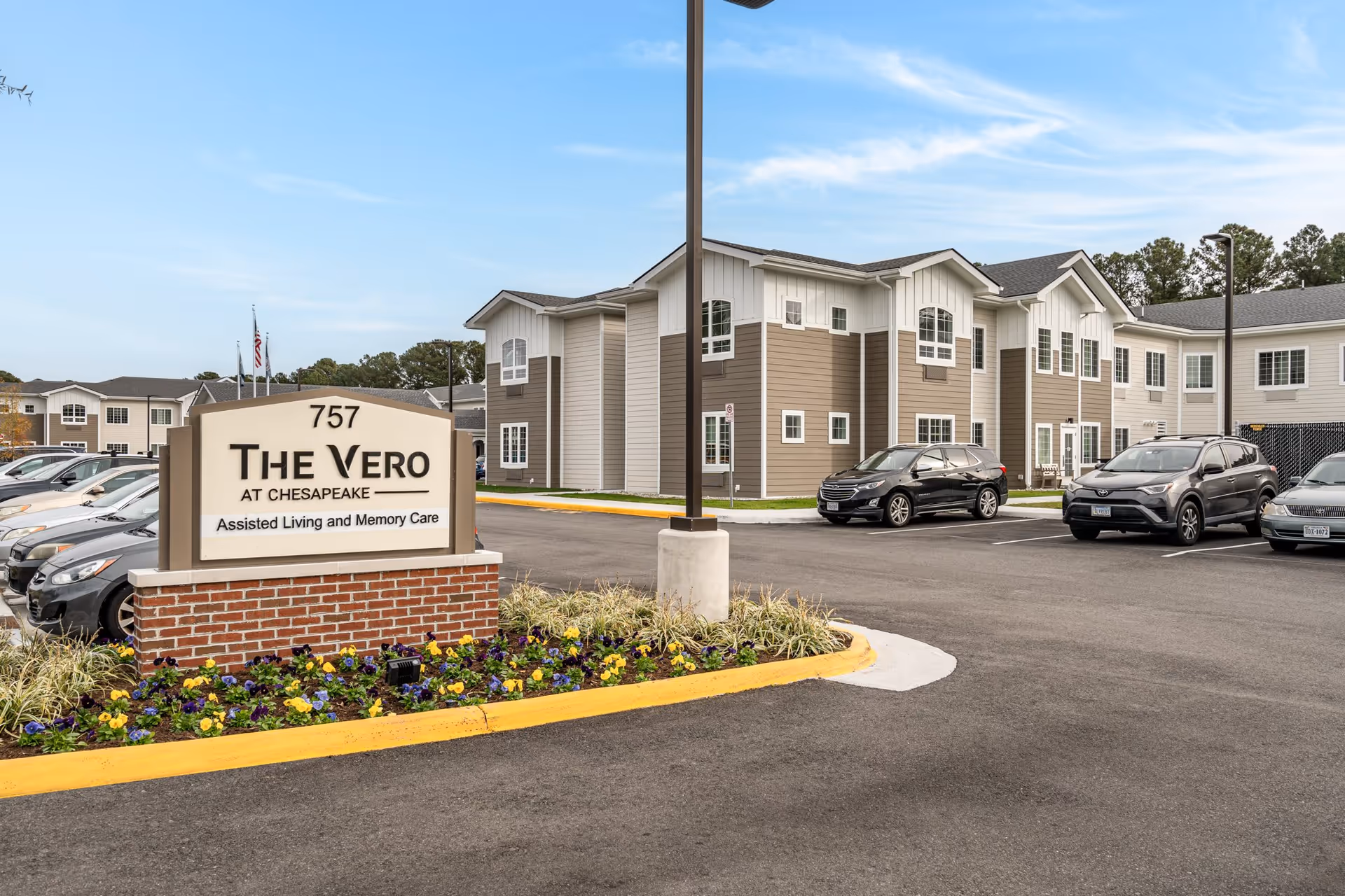 Exterior view of The Vero at Chesapeake Senior Living facility showing a parking lot with several cars, a sign with the facility name and address, and a two-story building with beige and white siding under a blue sky.