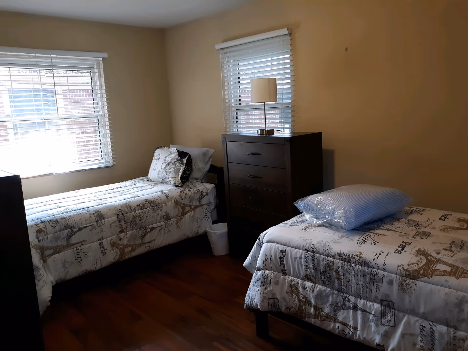 A bedroom with two single beds, each with white and beige bedding featuring Eiffel Tower and Paris-themed prints. Between the beds is a dark wooden dresser with a small lamp on top. The room has two windows with white blinds and wooden flooring.