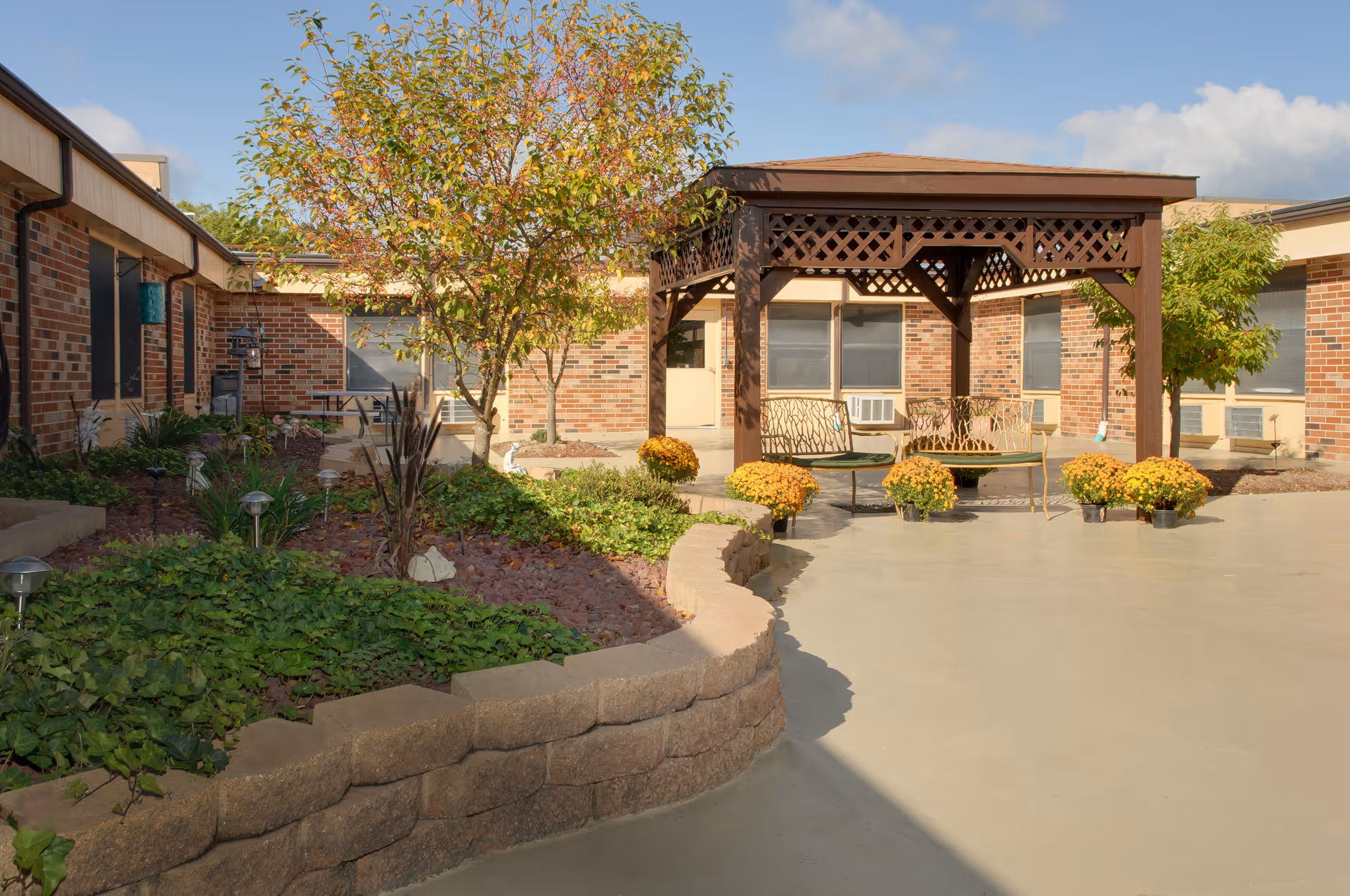 Outdoor courtyard area at a senior living facility with a wooden gazebo, metal benches, potted yellow flowers, a tree with autumn leaves, and a garden bed bordered by a low stone wall. The building surrounding the courtyard has brick walls and several windows.