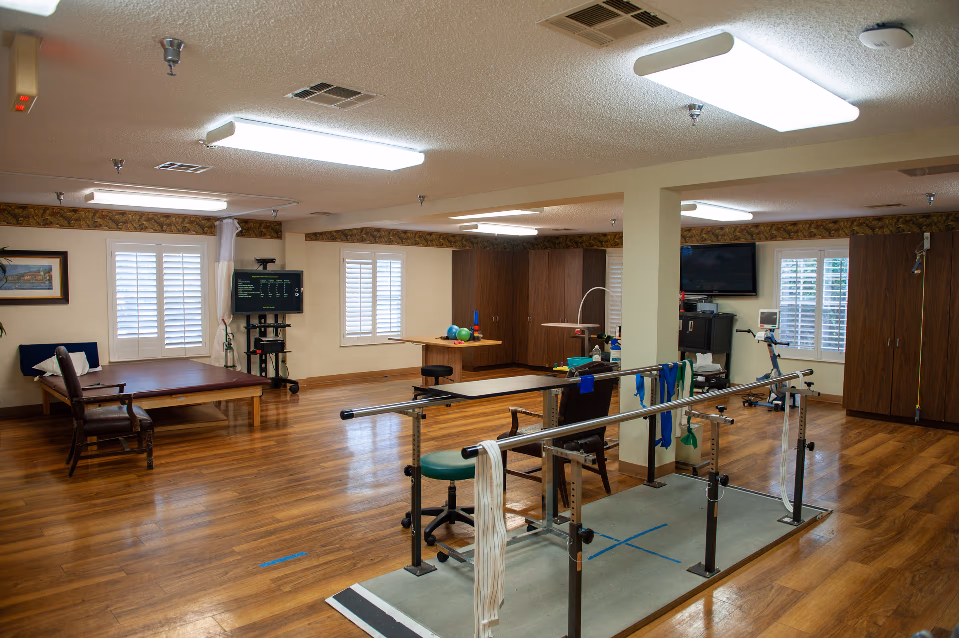 Spacious indoor therapy/recreation room with parallel bars, exercise equipment, tables, and wood floors.