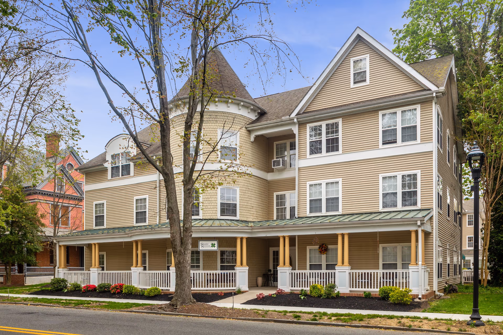 Exterior view of a three-story beige assisted living facility with white trim and a green metal roof over the porch. The building has multiple windows and a turret-like rounded section. There is a tree in front and a streetlamp on the right side. The porch is decorated with plants and a wreath, and a sign reads 'State Street Assisted Living.'