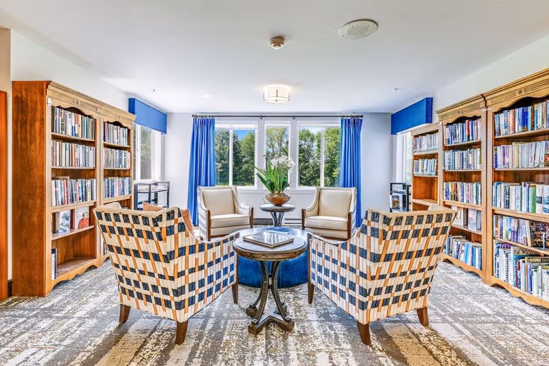 Bright senior living library lounge with bookshelves, four patterned armchairs arranged around a round table and a window with blue curtains.