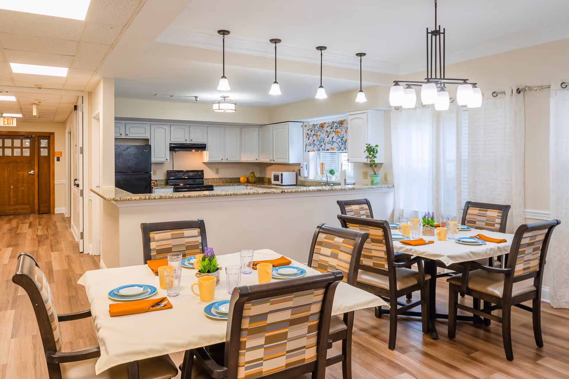 A bright dining area in a senior living facility with two tables set for meals, each with four chairs featuring patterned upholstery. The tables have white tablecloths, blue plates, orange napkins, yellow mugs, and clear glasses. Behind the dining area is a kitchen with white cabinets, a black refrigerator, stove, microwave, and a window with floral curtains. The floor is wood, and there are multiple ceiling lights illuminating the space. A wooden door and hallway are visible in the background.