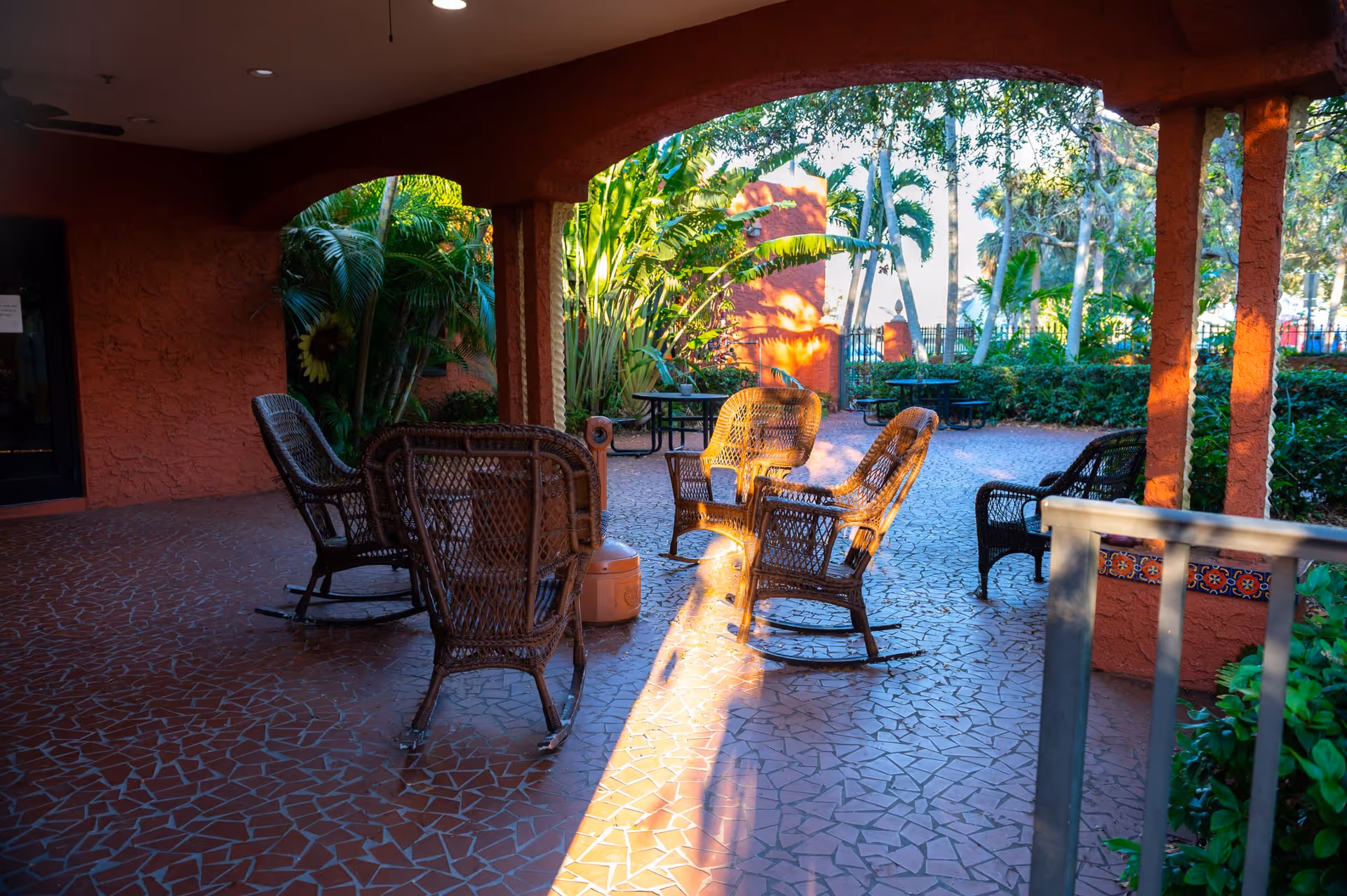 Outdoor covered patio area with several wicker rocking chairs arranged in a circle on a tiled floor. The patio is surrounded by lush green plants and trees, with sunlight casting shadows on the floor. There are also picnic tables visible in the background.