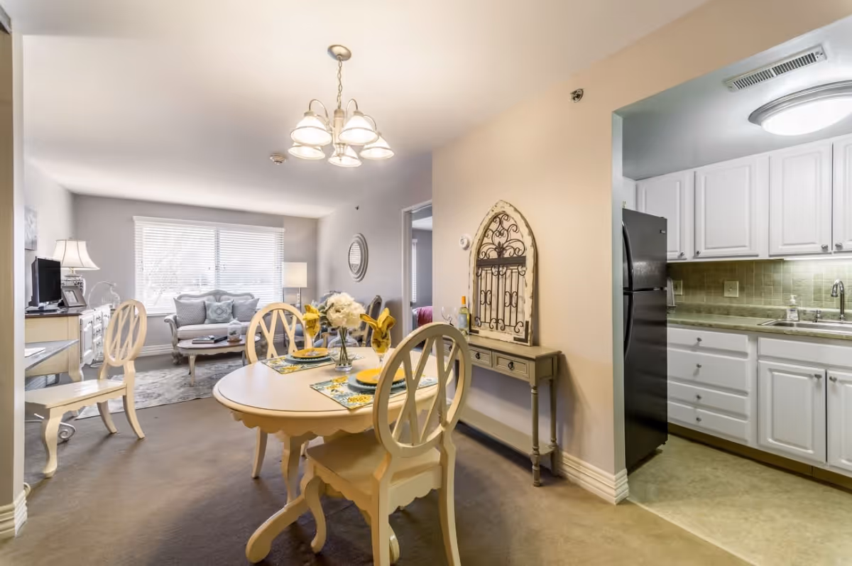 A bright and cozy senior apartment interior featuring a dining area with a round table set for four, a living room with a sofa and coffee table near a large window, and a kitchen with white cabinets and a black refrigerator.