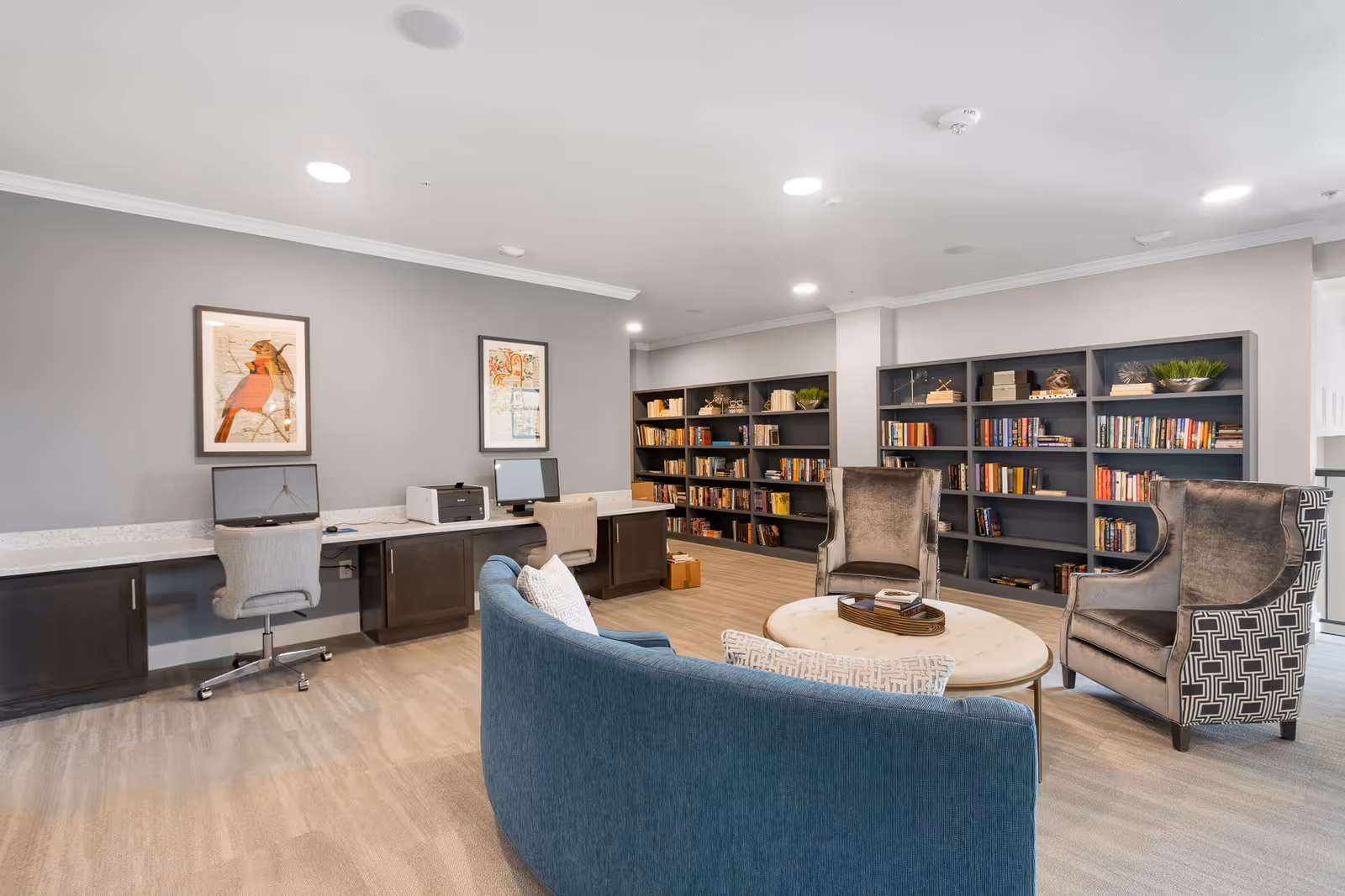 A cozy common area in a senior living facility featuring a curved blue sofa with patterned pillows, two upholstered armchairs, a round coffee table with books, built-in bookshelves filled with books and decorative items, and a long desk with two computer workstations and a printer. The walls are painted gray with framed artwork, and the floor has light wood-style flooring.
