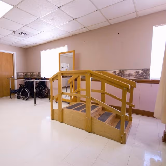 An indoor rehabilitation room with a wooden ramp and stairs for physical therapy. There are parallel metal bars on one side and two wheelchairs positioned near a closed wooden door. The room has beige walls with a decorative border and two windows letting in natural light.