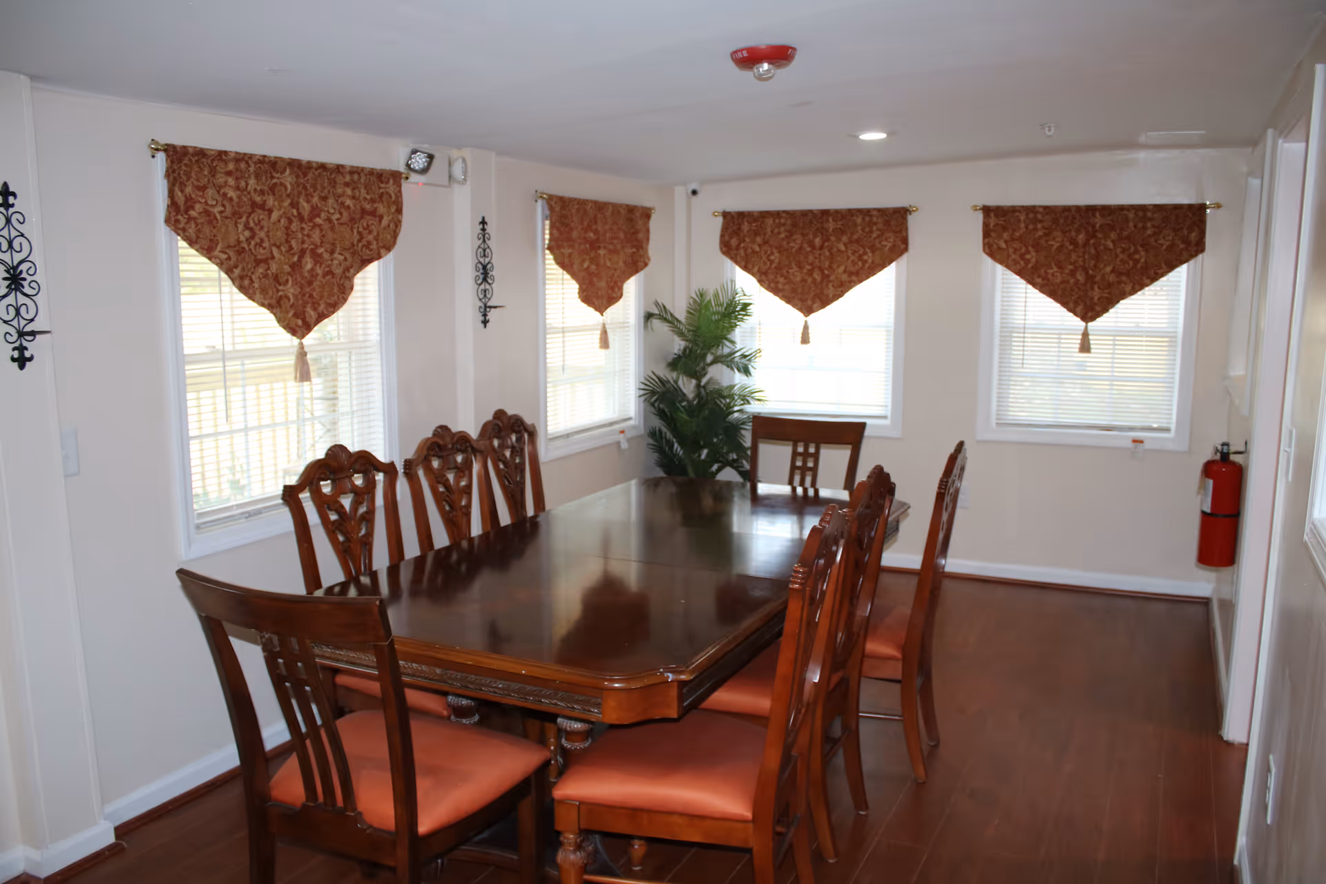 A dining room with a large polished wooden table surrounded by eight wooden chairs with red cushions. The room has four windows with red patterned valances and white blinds. There is a green potted plant in the corner and a red fire extinguisher mounted on the wall. The floor is wooden and the walls are painted white.