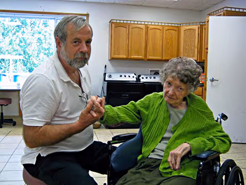 An elderly woman in a wheelchair wearing a green cardigan is holding hands with a man in a white polo shirt inside a room with wooden cabinets and a stove in the background.