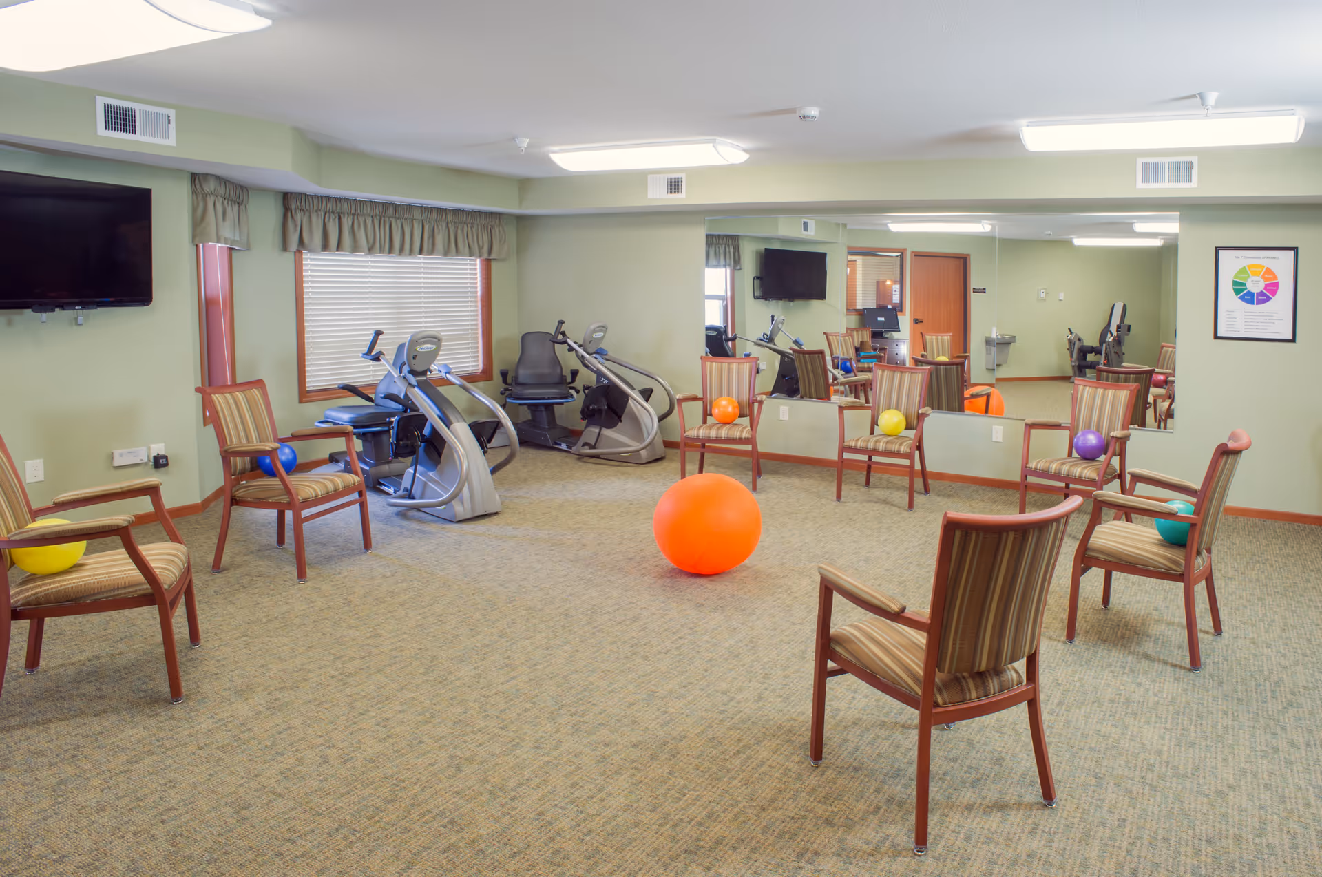 A senior living facility exercise room with several striped wooden chairs arranged in a circle, each holding a small colorful exercise ball. In the center of the room is a large orange exercise ball. The room has light green walls, a large mirror on one side, two exercise machines near a window with blinds, a wall-mounted TV, and a framed colorful chart on the wall.