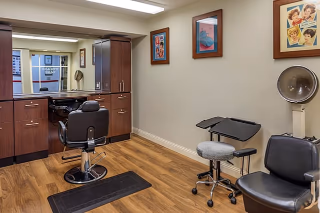Interior of a salon room with wooden flooring, a large mirror above a wooden cabinet, a black salon chair in front of the mirror, a rolling stool, a black chair, and a vintage hair dryer. The walls are decorated with framed pictures.