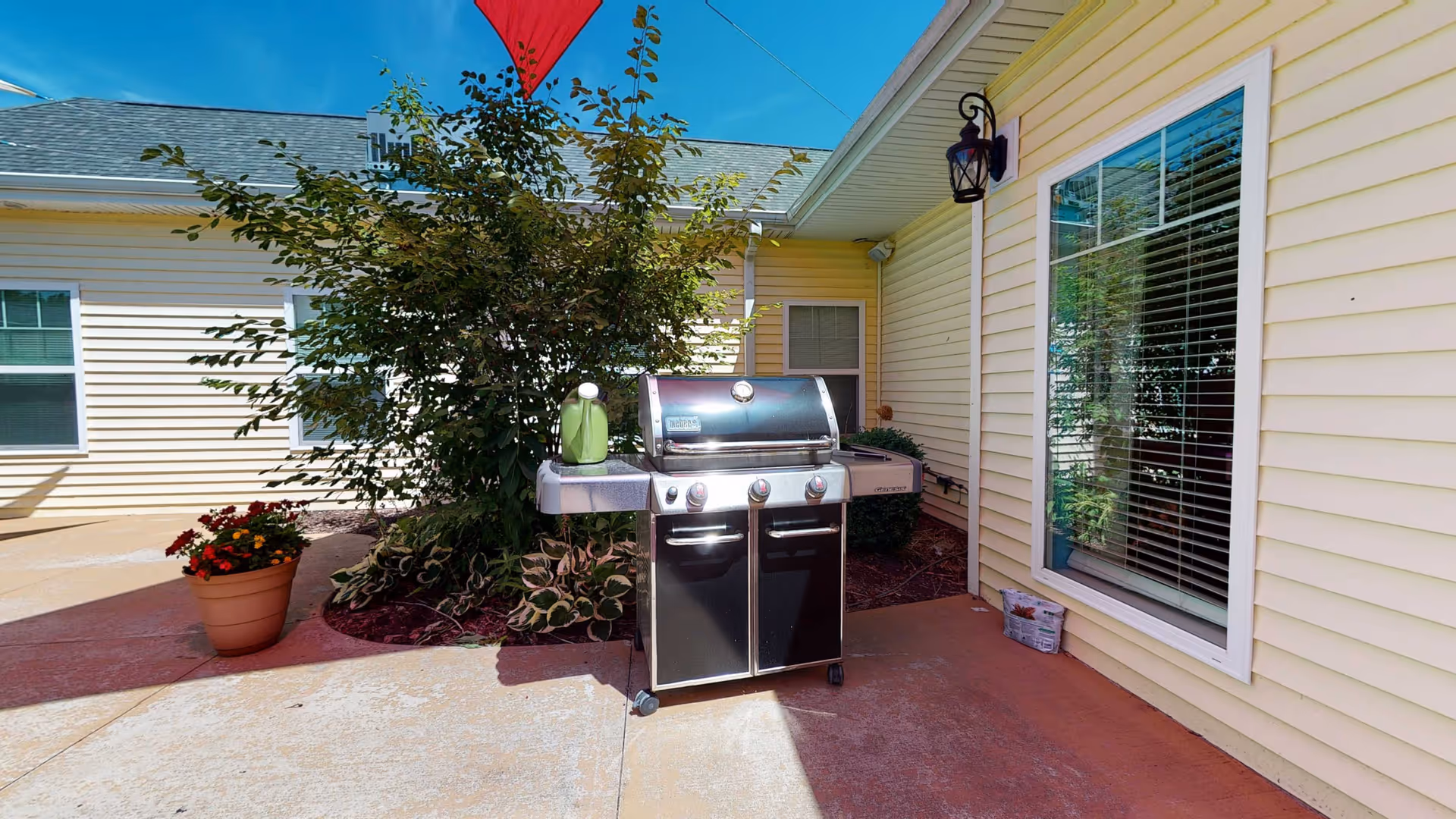 Outdoor patio area with a black gas grill on wheels placed on a concrete surface. There is a potted plant with red and yellow flowers to the left and a leafy green shrub behind the grill. The building exterior is light yellow with white trim, featuring a large window with blinds and a wall-mounted lantern light fixture. A red umbrella is partially visible at the top of the image.