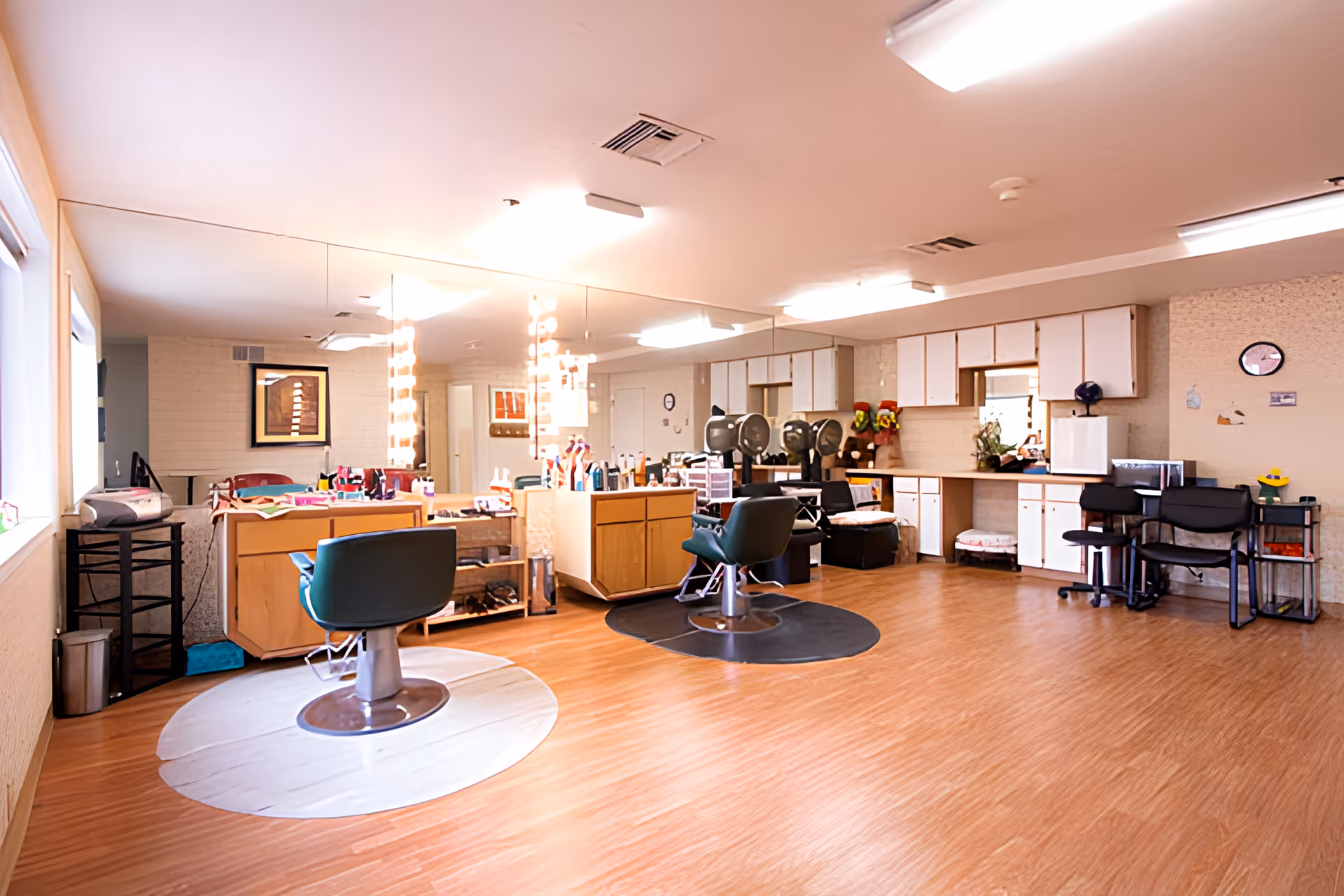 Interior of a salon area in a senior living facility with two styling chairs in front of large mirrors, hair care products on counters, hair dryers, and additional seating along the wall. The room has wooden flooring and bright overhead lighting.