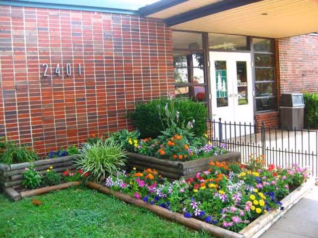 Brick facility entrance with glass doors and a colorful flower garden in front.