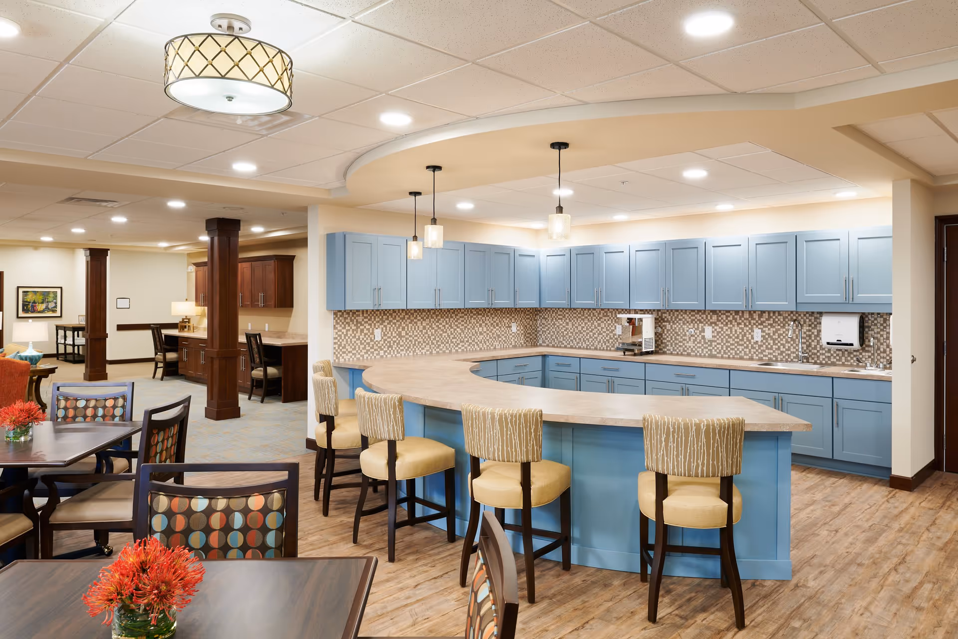 A bright and spacious kitchen area with light blue cabinets and a curved countertop with four cushioned bar stools. The kitchen features a mosaic tile backsplash, pendant lights hanging from the ceiling, and a coffee machine on the counter. Adjacent to the kitchen are dining tables with chairs that have colorful patterned cushions. The room has wood flooring and recessed lighting, with a cozy and welcoming atmosphere.