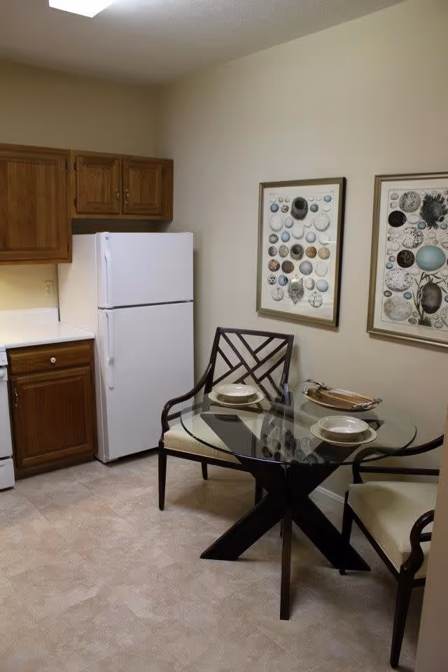 A small kitchen area with wooden cabinets, a white refrigerator, and a white stove. Next to the kitchen is a round glass dining table with two cushioned chairs. The table is set with two plates and a decorative tray. Two framed artworks featuring various circular patterns hang on the beige wall behind the table.