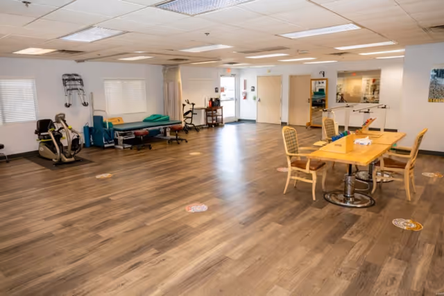 A spacious room with wooden flooring featuring exercise equipment such as a stationary bike and therapy tables on the left side, and a wooden table with chairs on the right side. The room has white walls, windows with blinds, ceiling lights, and mirrors on the far wall.