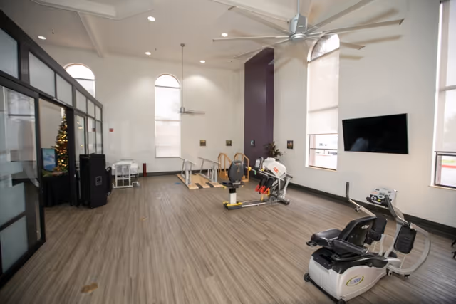 Large bright exercise room with senior fitness equipment, wood-look flooring, tall arched windows, and a wall-mounted TV.