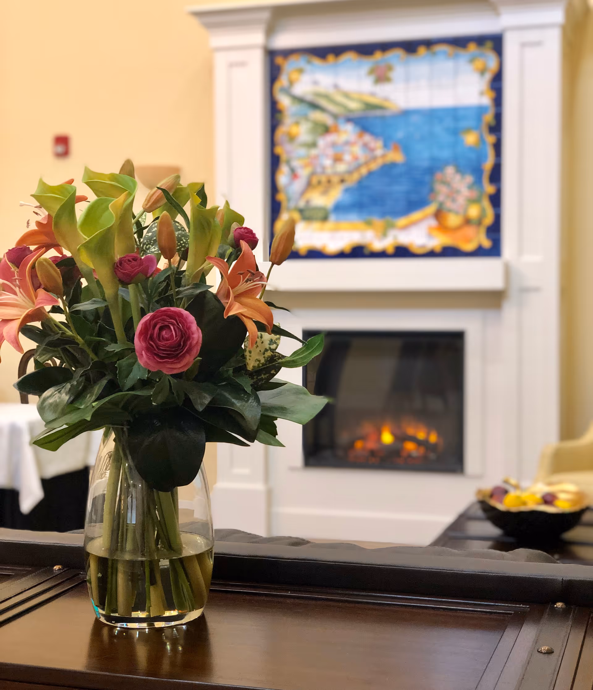 A clear glass vase filled with a bouquet of flowers including pink roses, orange lilies, and green calla lilies sits on a wooden table. In the background, there is a white fireplace with a colorful tile mural above it depicting a coastal scene, and a bowl of fruit on a nearby surface.
