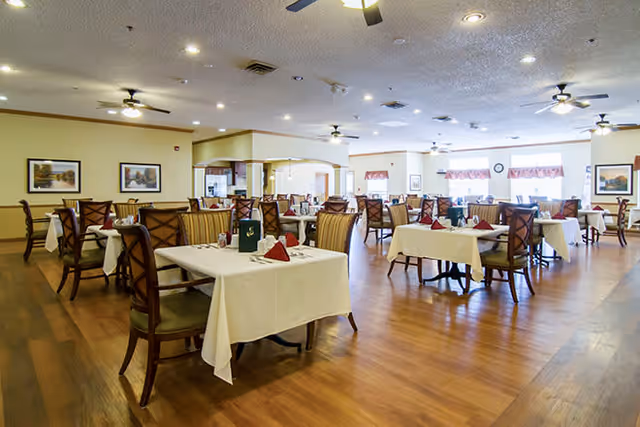 A spacious dining room with multiple tables covered in white tablecloths, each set with napkins, glasses, and menus. The room has wooden floors, ceiling fans, and framed artwork on the walls. Large windows allow natural light to fill the space.