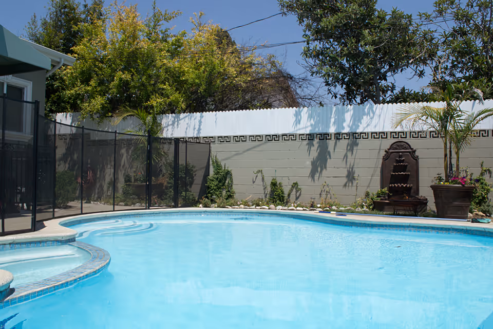 Outdoor swimming pool with clear blue water surrounded by a safety fence. There are plants and trees around the pool area, a decorative wall fountain, and a white fence in the background under a clear blue sky.