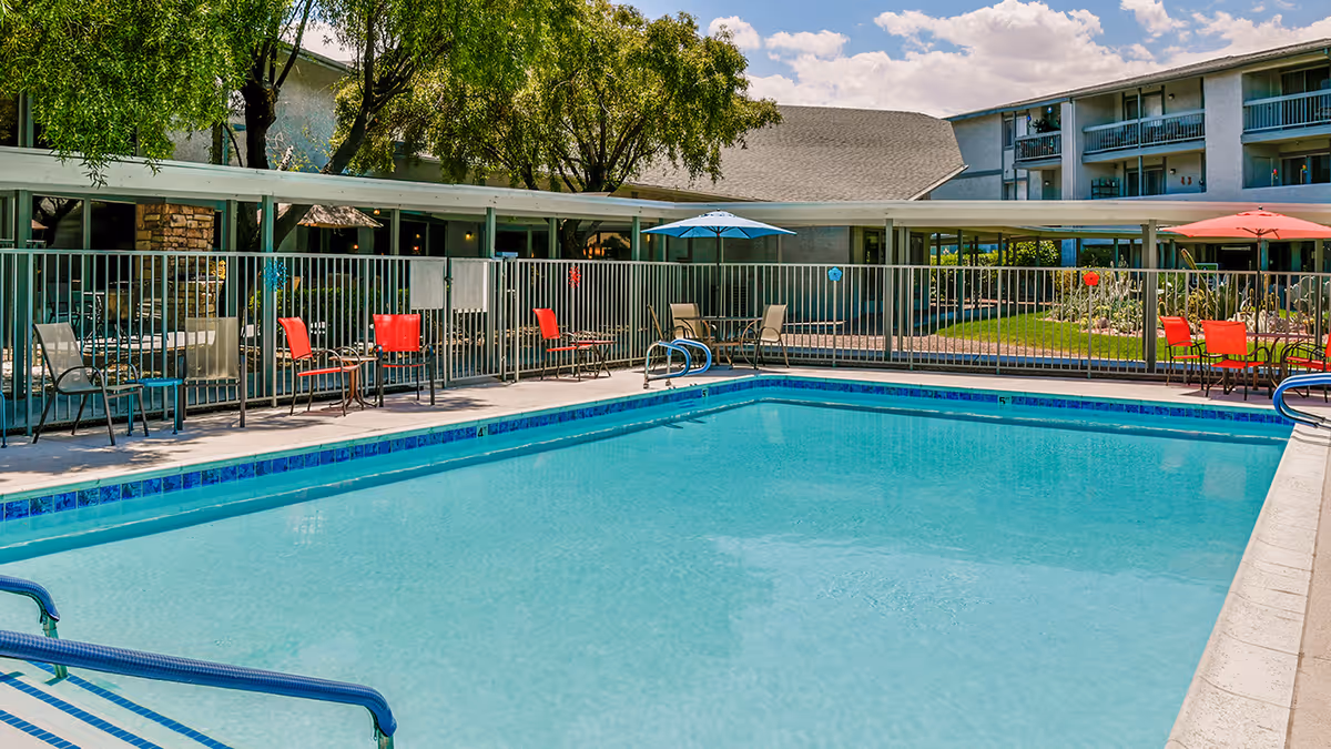 Outdoor swimming pool area at Holiday Montara Meadows with clear blue water, surrounded by a safety fence. Several chairs and tables with umbrellas are placed around the pool. Trees and a multi-story building are visible in the background under a partly cloudy sky.