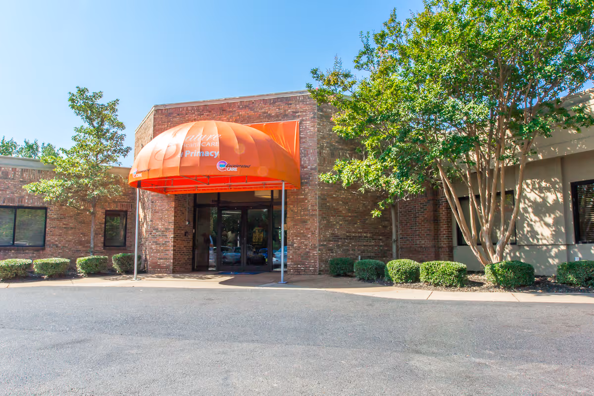 Entrance of a brick building with an orange awning that reads 'Signature HealthCARE of Primacy'. The entrance has glass double doors and is flanked by small bushes and trees under a clear blue sky.
