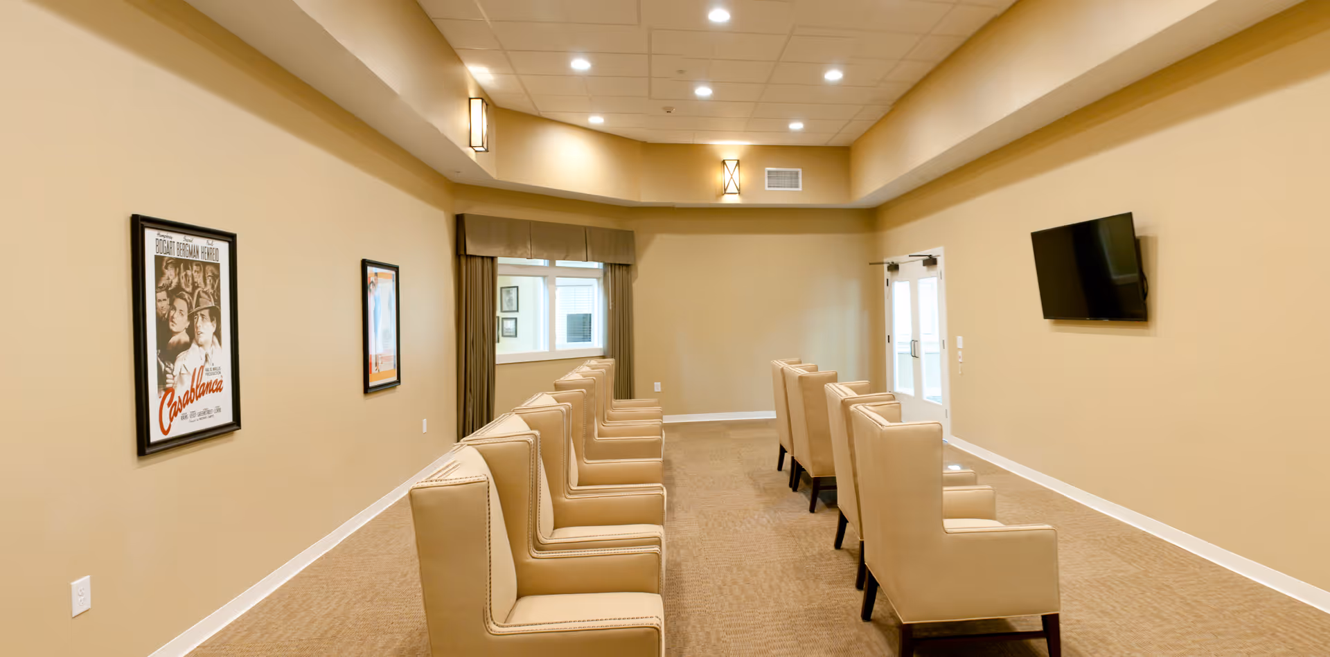 A quiet room with two rows of beige armchairs facing a wall-mounted flat screen TV. The walls are painted light beige and decorated with framed movie posters. There is a window with curtains and a double door at the far end of the room. The ceiling has recessed lighting and wall sconces.