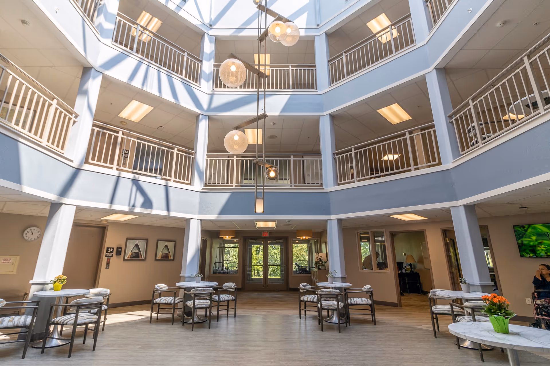 Interior view of a senior living facility with a spacious, multi-level atrium featuring railings on the upper floors. The ground floor has several small round tables with chairs arranged around them, some with potted plants on top. Large windows and glass doors at the far end allow natural light to enter, complemented by ceiling lights and hanging spherical pendant lamps. A person in a wheelchair is visible on the right side near a television mounted on the wall.