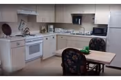 A kitchen area with white cabinets, a white stove with a kettle on it, a microwave, a refrigerator, and a small wooden table with two floral upholstered chairs. There are some kitchen items on the countertops including a toaster and a plant.
