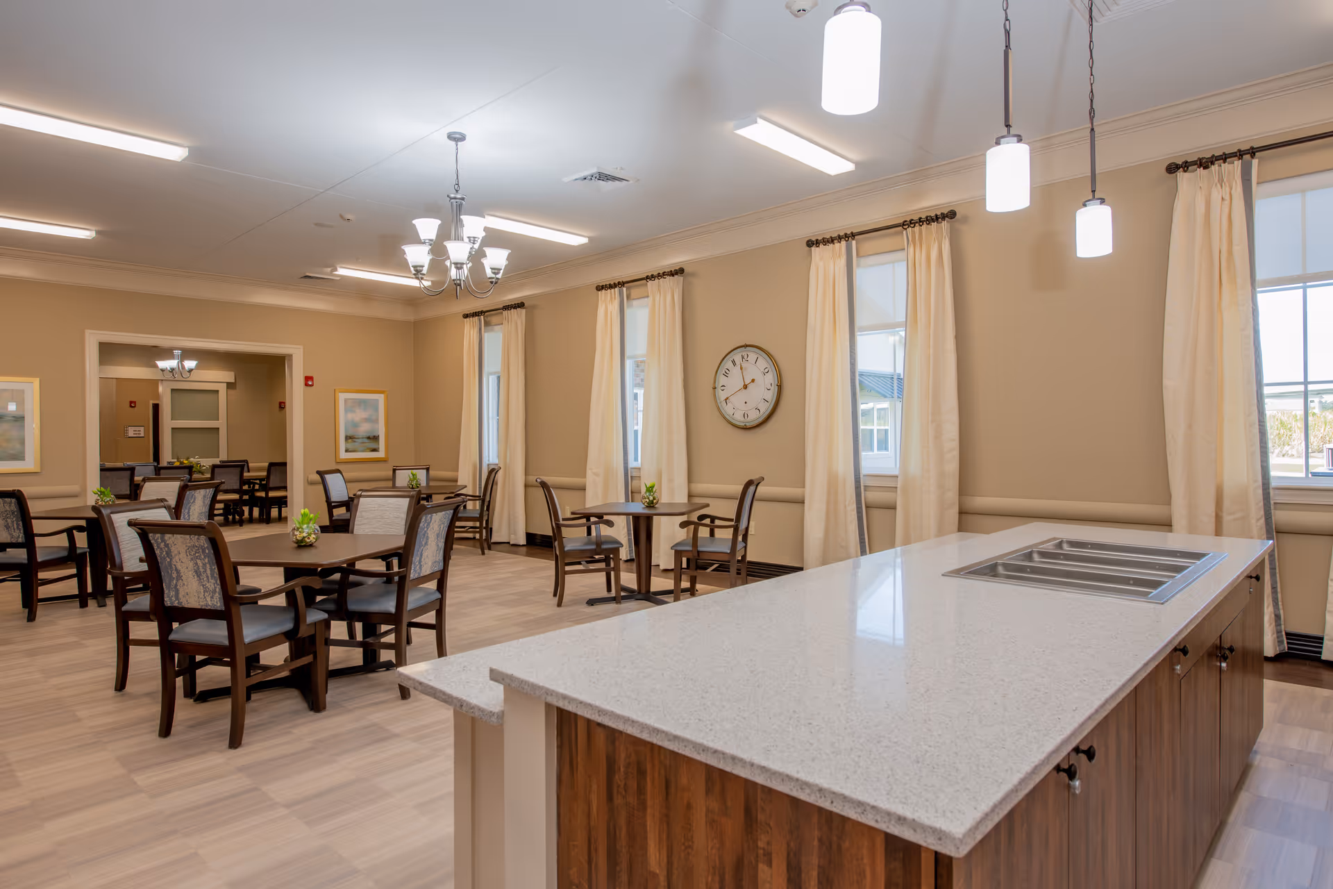 A spacious dining room in an assisted living facility with multiple wooden tables and chairs arranged neatly. The room features beige walls, large windows with cream curtains, a wall clock, and a central island with a light-colored countertop and built-in sink. Overhead lighting includes pendant lights and a chandelier.