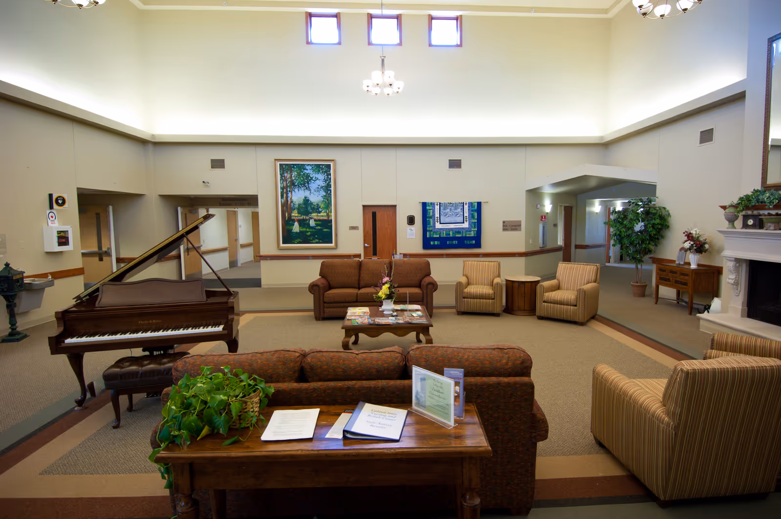 A spacious and well-lit common area in a nursing and rehab center featuring comfortable seating including sofas and armchairs arranged around a coffee table. There is a grand piano on the left side and a fireplace on the right side with decorative plants and flowers. The walls are adorned with framed artwork and the ceiling has small windows near the top allowing natural light in.