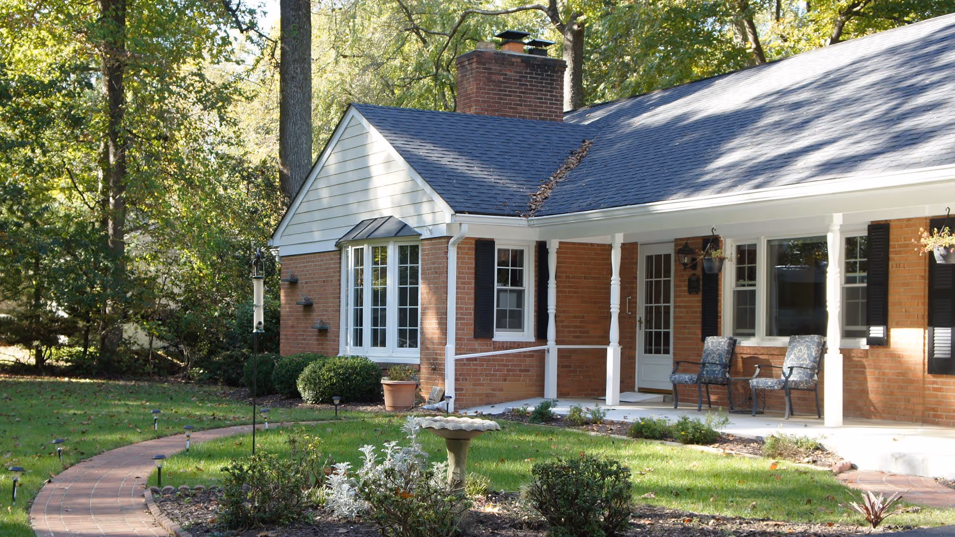 Exterior view of a single-story brick house with a dark shingled roof, white columns supporting a covered porch, two chairs on the porch, a birdbath in the front yard, and a curved brick pathway surrounded by green grass and trees.