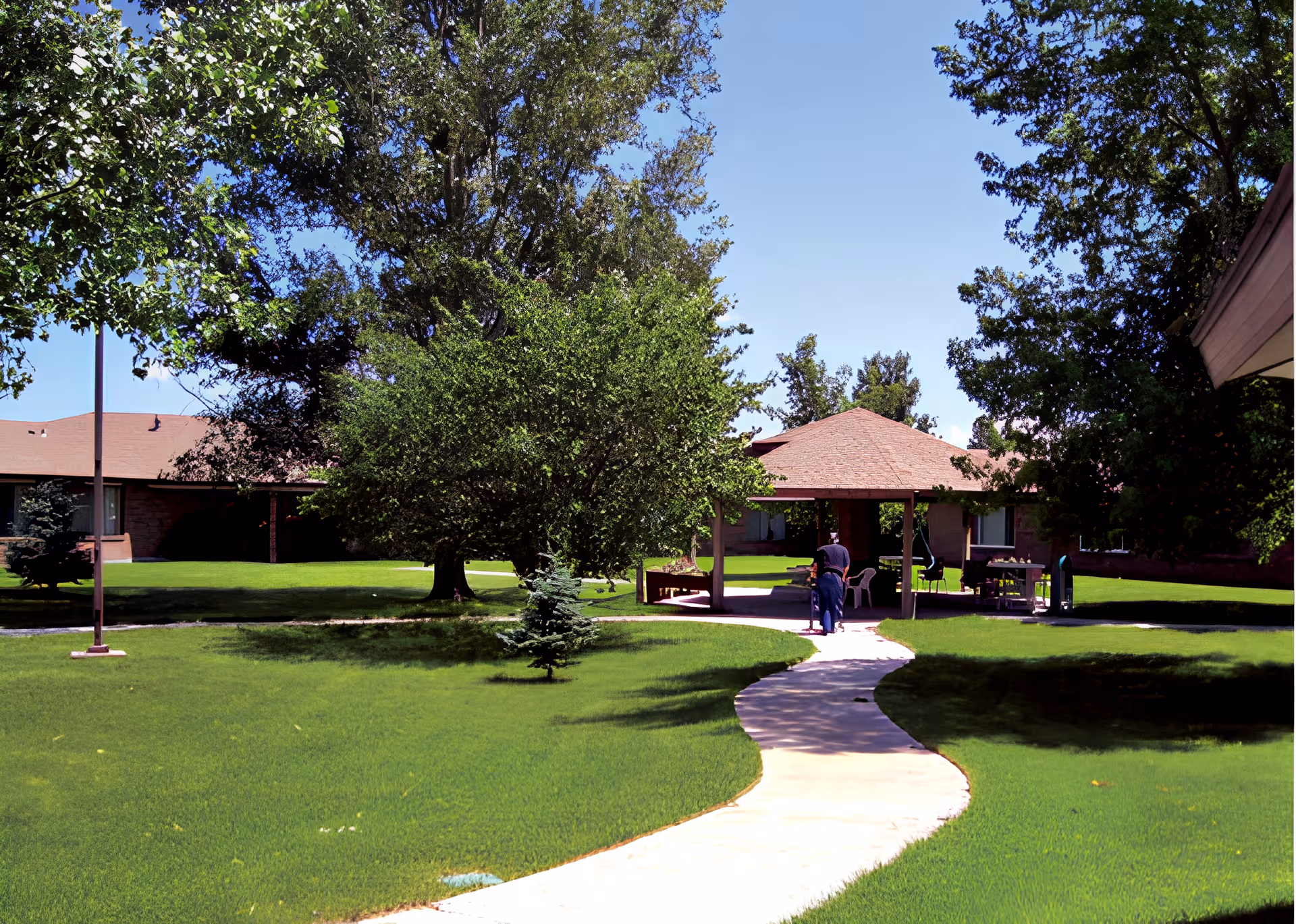 A winding concrete pathway leads through a well-maintained grassy courtyard with large trees and a gazebo. A person using a walker is seen walking towards the gazebo, which has chairs and tables underneath. Surrounding the courtyard are single-story buildings with brown roofs under a clear blue sky.