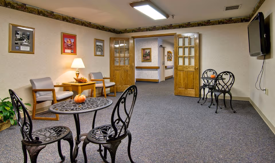 A common area in a senior living facility with two small round black metal tables, each with two matching chairs. One table has a small pumpkin on it. There are two beige armchairs with wooden arms next to a wooden side table with a lamp and decorative items. The walls are light-colored with framed pictures and a floral border near the ceiling. Double wooden doors with glass panels lead to a hallway. A flat-screen TV is mounted on the right wall.