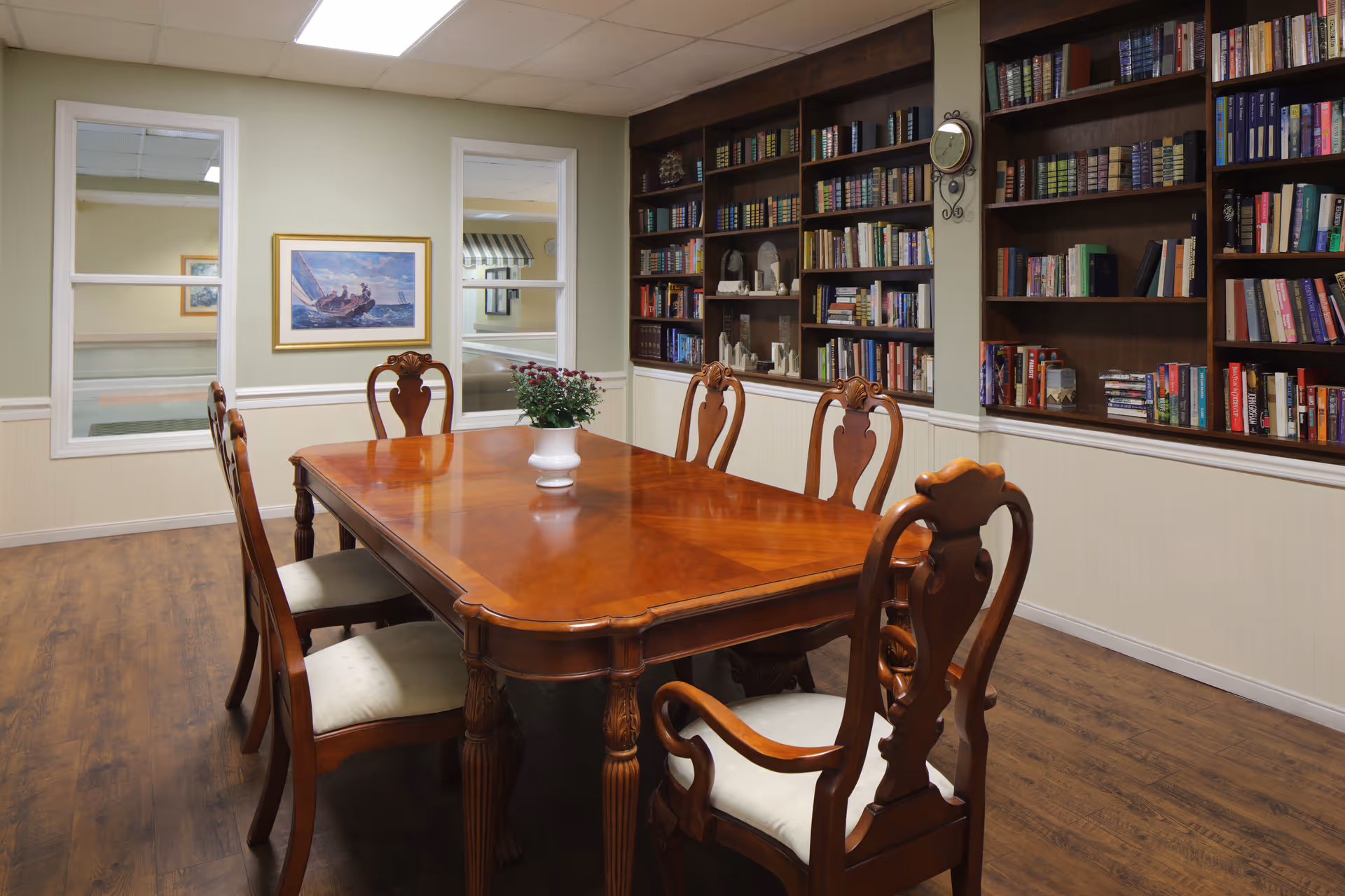 A cozy room with a polished wooden table surrounded by six wooden chairs with cushioned seats. The room features built-in dark wood bookshelves filled with books and decorative items, a wall clock, and two windows with white frames. A framed painting of a sailboat is hung on the wall, and a small potted plant is placed on the table. The floor is wooden, and the walls are painted light green with white wainscoting.