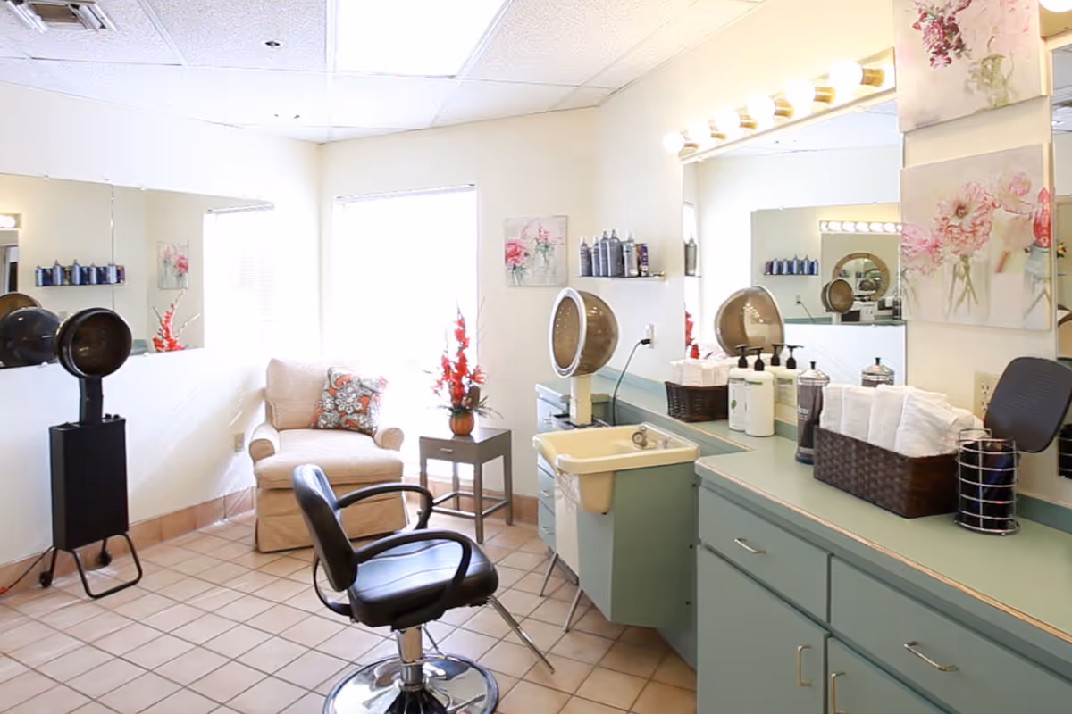 A bright and clean salon area with a black salon chair in front of a large mirror and a hair dryer hood. There is a beige armchair with a decorative pillow next to a small side table holding a vase with red flowers. The countertop has various hair care products, a basket with white towels, and a small sink. The walls are decorated with floral artwork.