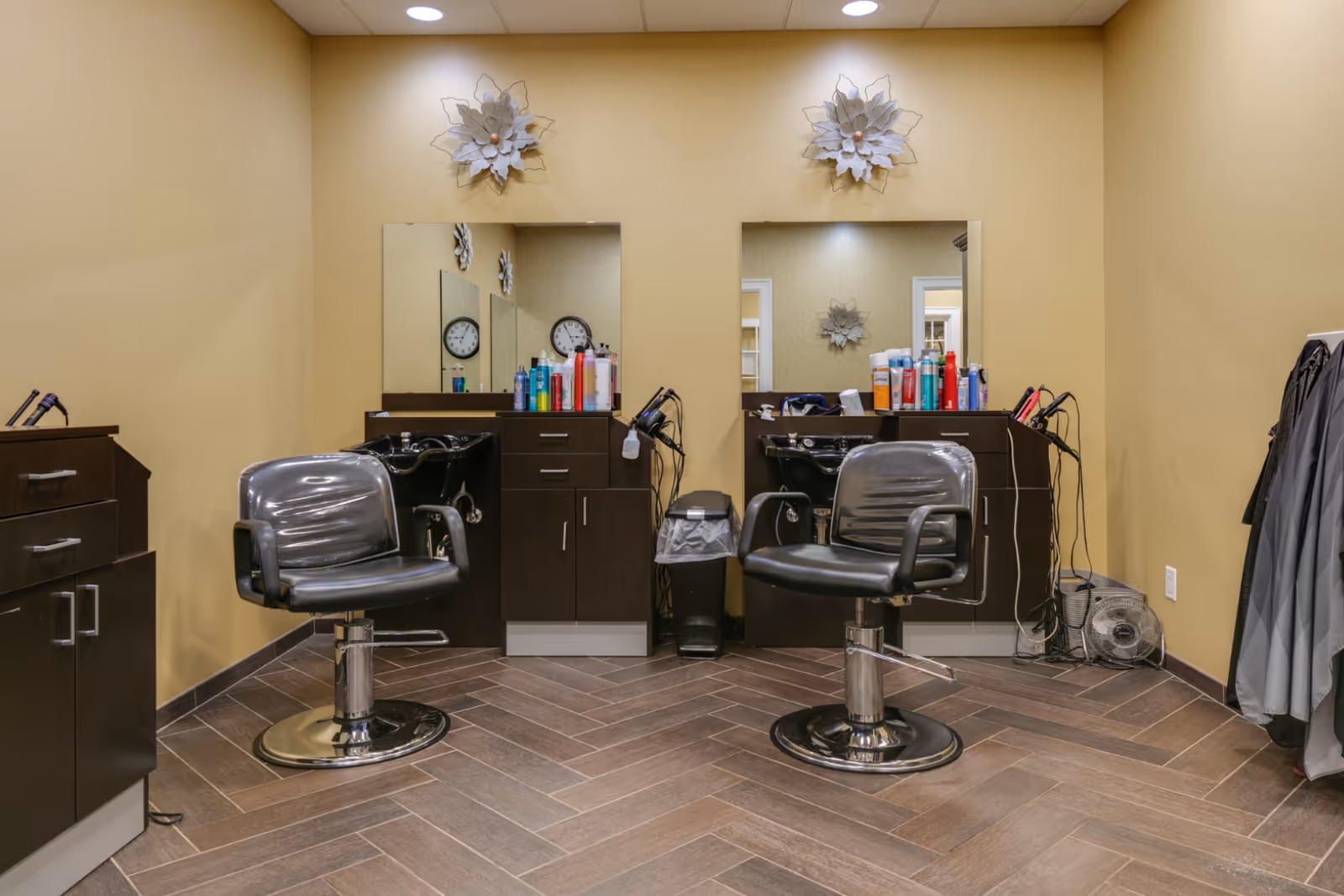 Interior view of a senior living facility's hair salon area with two black salon chairs in front of mirrors. The space includes dark wood cabinetry, hair care products on the counters, a small trash bin, and a fan on the floor. The walls are painted beige and decorated with two large white floral wall hangings above the mirrors.