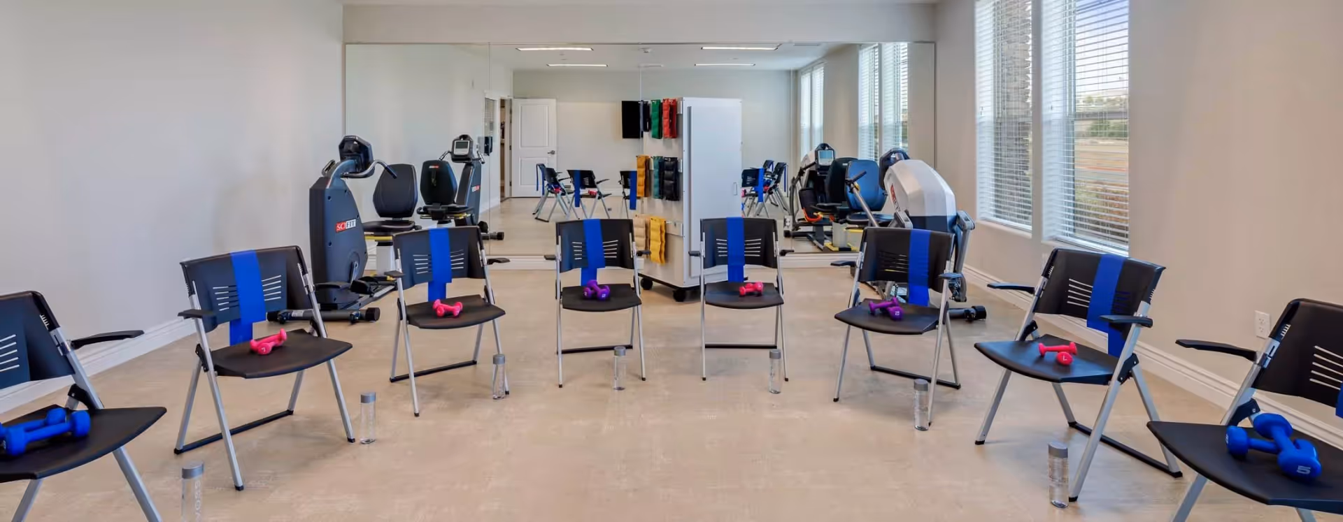 A fitness room with a circle of black chairs, each with a colored resistance band attached to the back and a pair of dumbbells on the seat. Water bottles are placed on the floor in front of each chair. The room has large windows on one side and a mirrored wall on the other, reflecting exercise equipment such as stationary bikes and resistance machines.