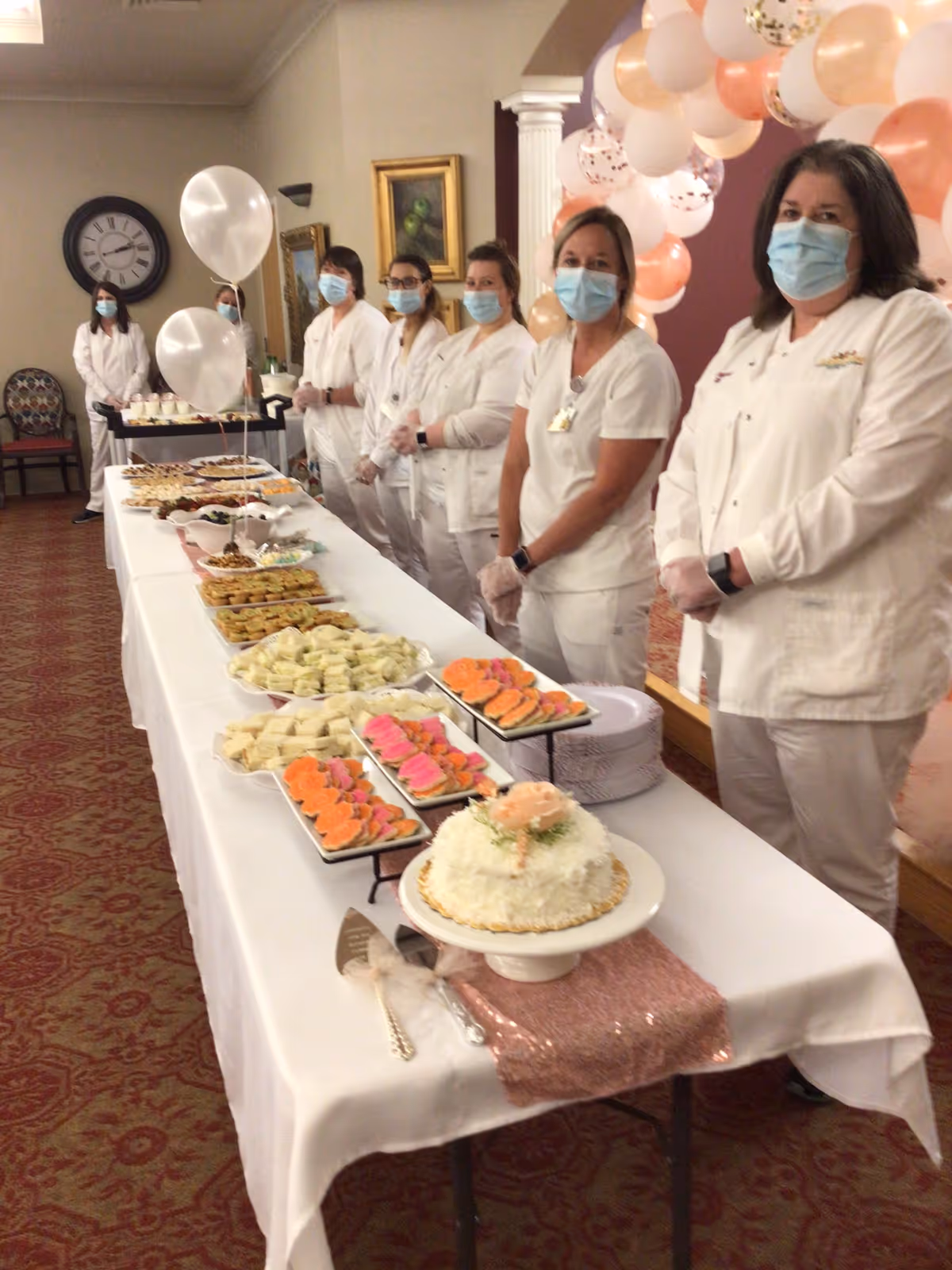 A group of seven healthcare workers wearing white uniforms, face masks, and gloves stand behind a long table covered with a white tablecloth. The table is filled with various trays of finger foods, sandwiches, cookies, and a decorated cake. There are white and peach balloons arranged in an arch in the background, and the setting appears to be an indoor room with carpeted flooring and framed artwork on the walls.