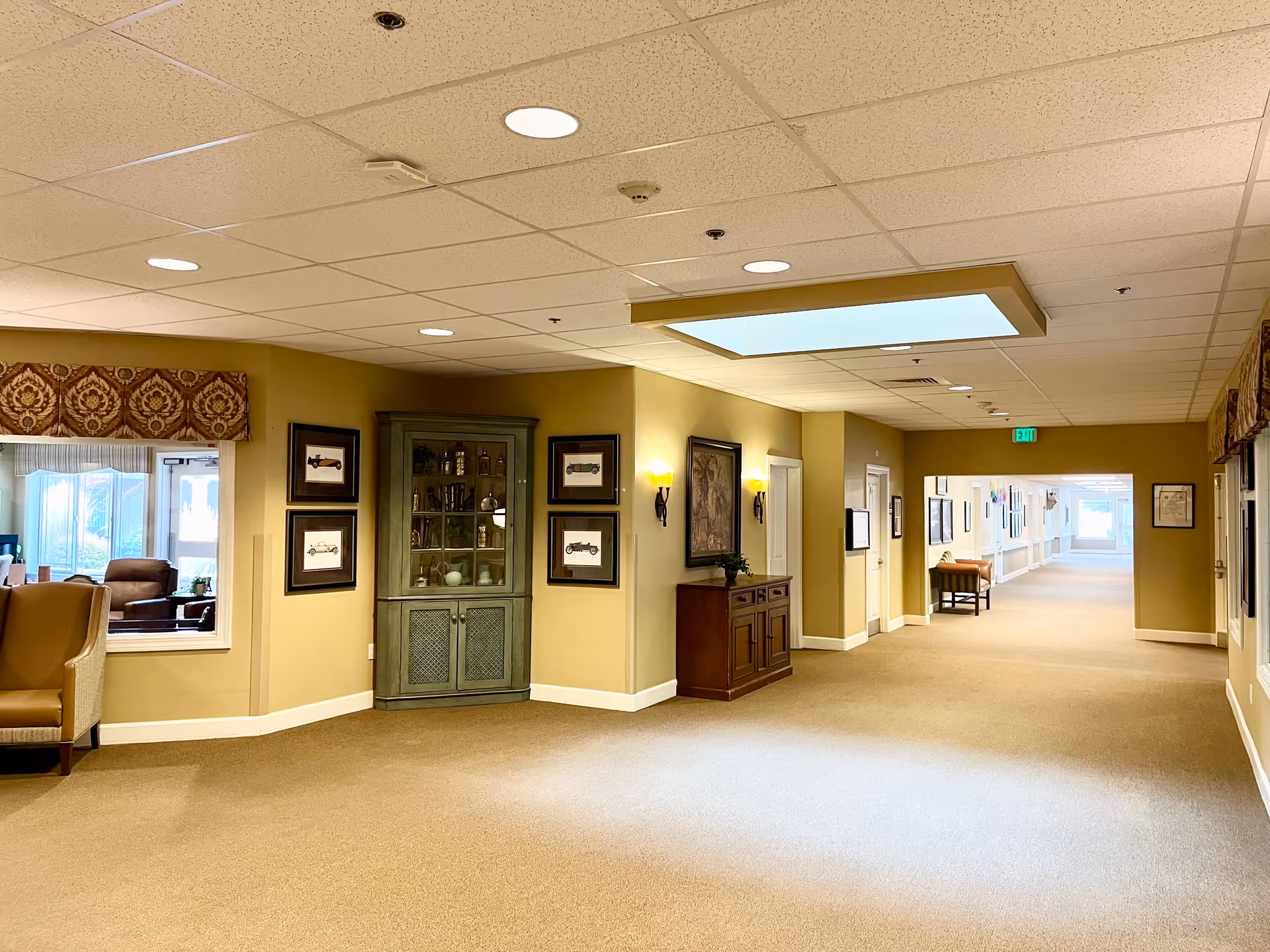 A wide, well-lit hallway in an assisted living facility with beige walls and carpeted floors. The hallway features framed artwork, a green display cabinet with glass doors, a wooden sideboard, and several chairs along the walls. There is a window on the left side showing a seating area with armchairs. The ceiling has recessed lighting and a large rectangular light fixture.