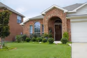 Front exterior view of a single-story brick house with an arched entryway, a dark wooden front door, a garage door on the right, and a well-maintained lawn with shrubs and bushes in front.
