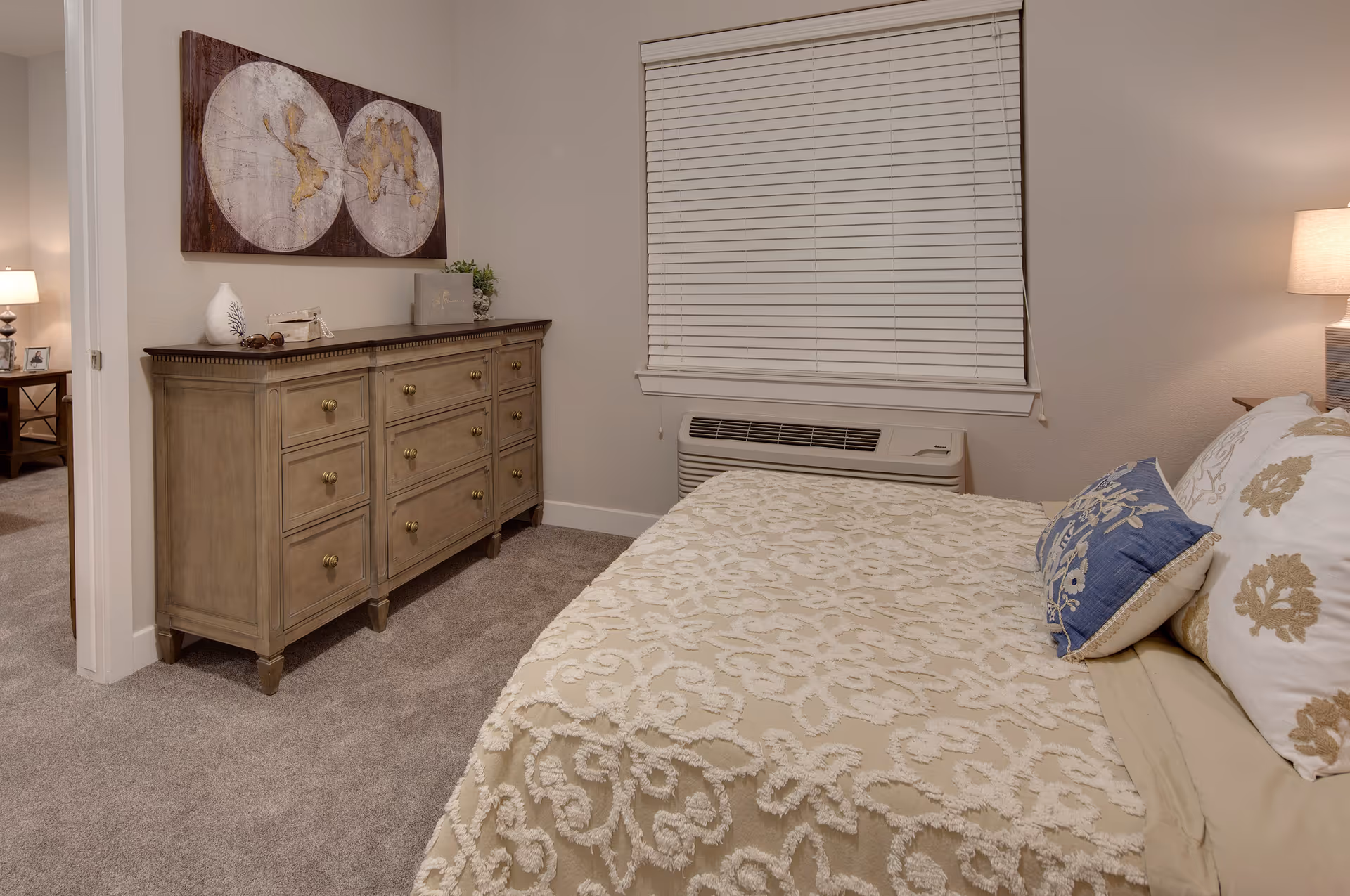 A cozy bedroom with a beige patterned bedspread and decorative pillows on the bed. There is a wooden dresser with brass knobs against the wall, topped with decorative items including a white vase and a small plant. Above the dresser hangs a world map artwork. A window with closed white blinds is above an air conditioning unit. The room has beige carpet and neutral-colored walls, with a glimpse of another room visible through an open doorway.
