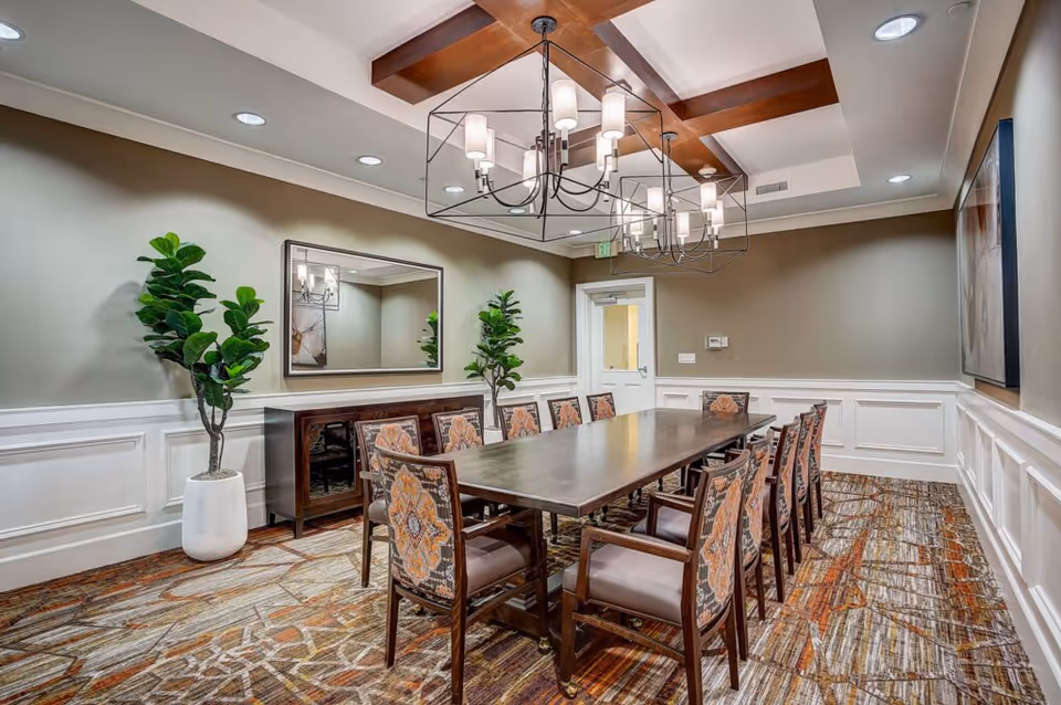 A well-lit dining room with a long rectangular wooden table surrounded by twelve upholstered chairs with patterned backs. The room features a decorative ceiling with wooden beams and two modern chandeliers. There are two large potted plants, a mirror on one wall, and a sideboard beneath the mirror. The walls are painted beige with white wainscoting, and the carpet has a multicolored abstract pattern.