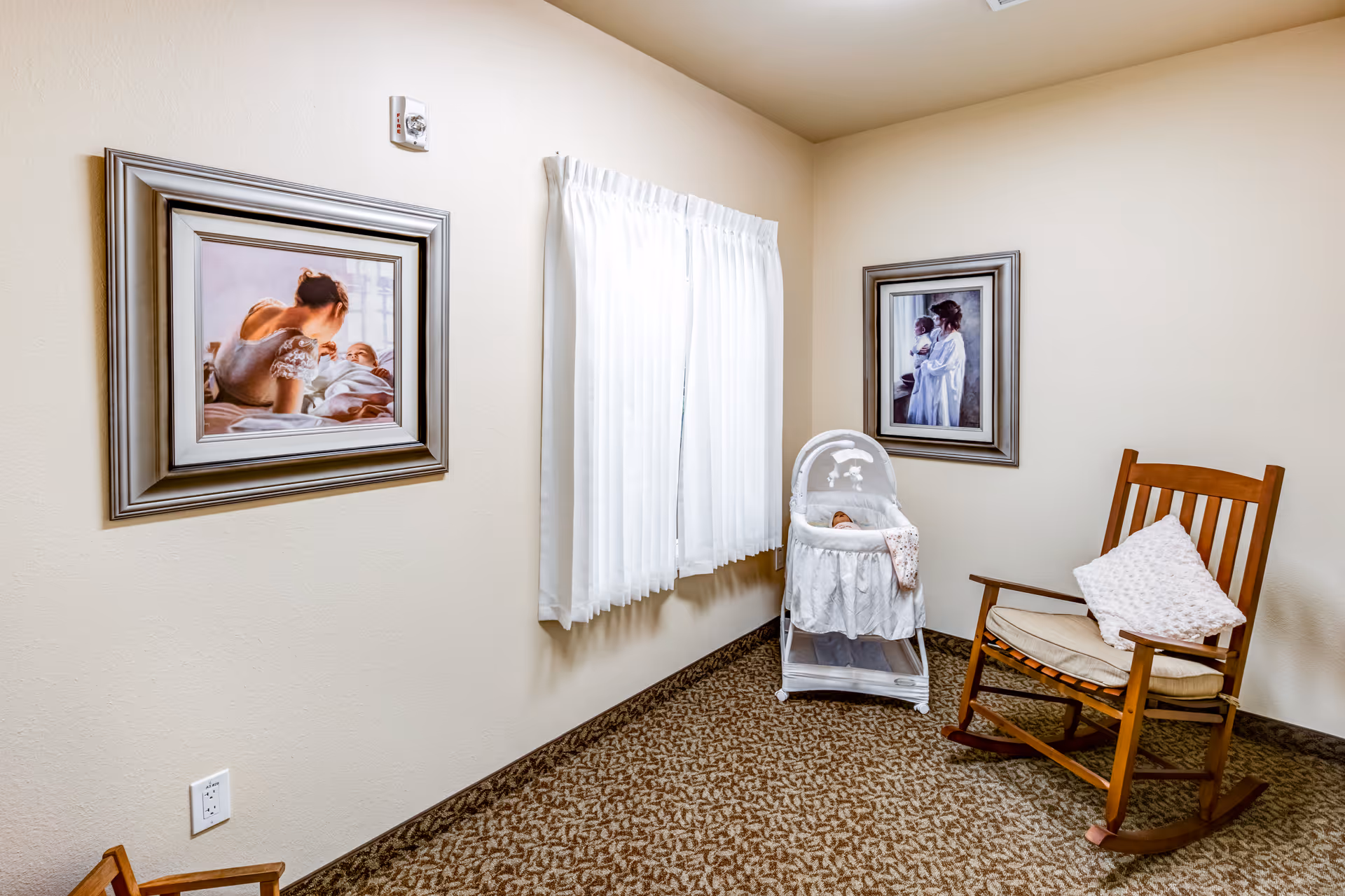 A cozy corner of a room with beige walls and patterned carpet. There is a wooden rocking chair with a white cushion and pillow, a white bassinet with a baby doll inside, and two framed pictures on the walls depicting a mother and child. A window with white curtains lets in natural light.