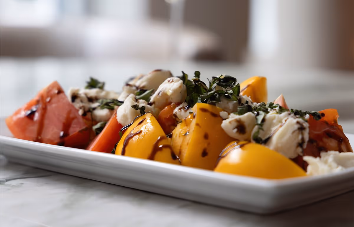 Close-up of a fresh tomato and mozzarella salad drizzled with balsamic glaze and garnished with chopped basil, served on a rectangular white plate.