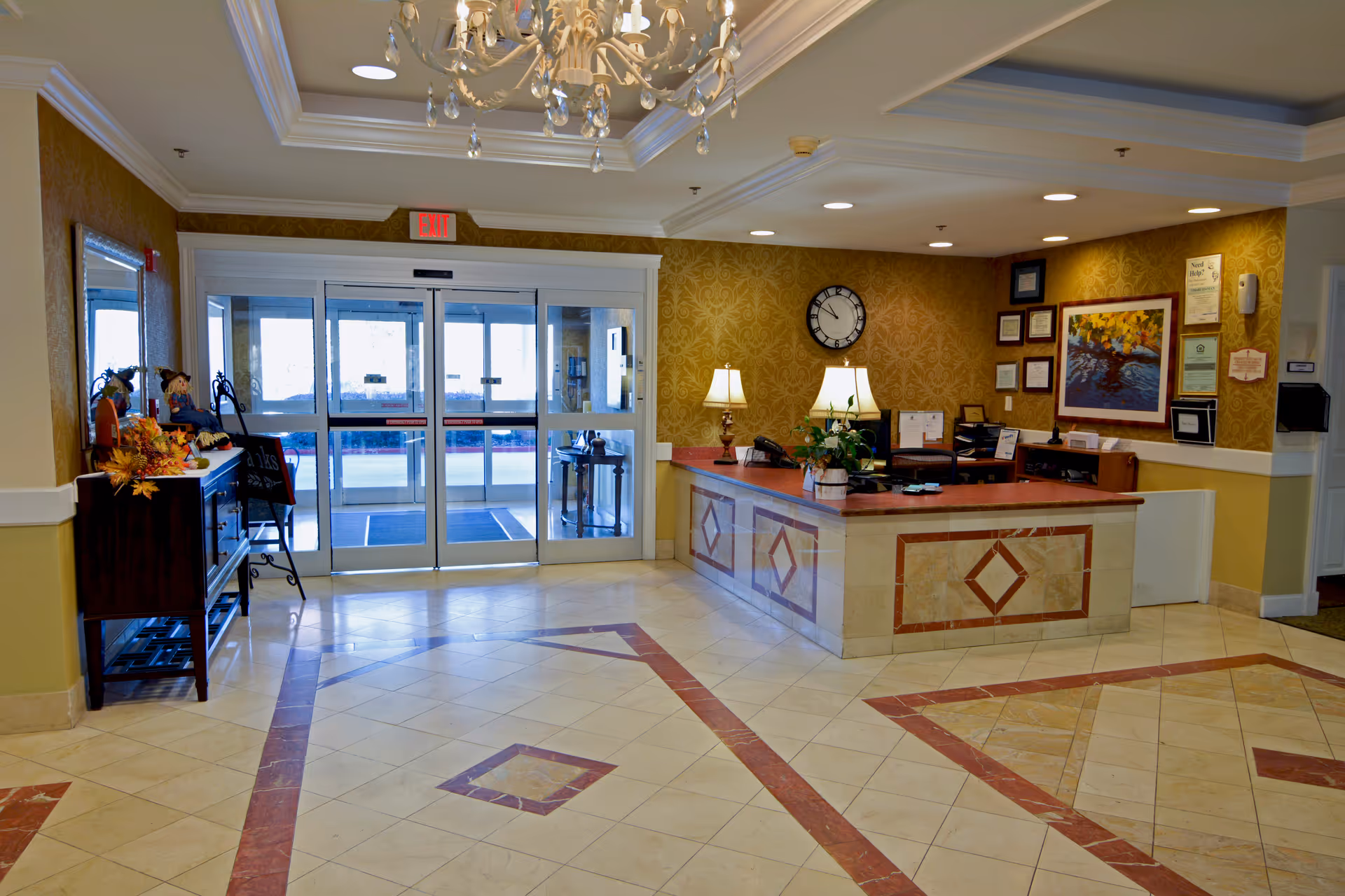 Reception area of Magnolia Place of Roswell featuring a front desk with two lamps, a clock on the wall, framed certificates and artwork, a chandelier hanging from the ceiling, and glass automatic entrance doors with an exit sign above. The floor has a decorative tile pattern and there is a small table with fall-themed decorations on the left side.