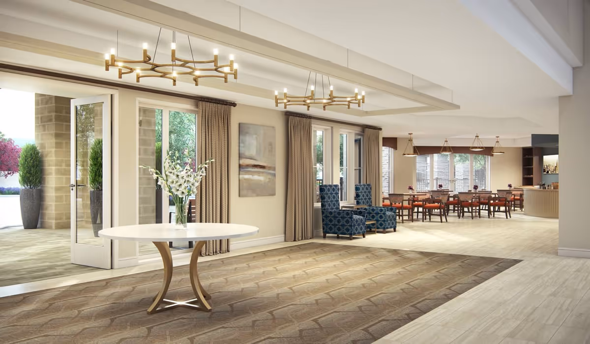 Spacious senior living common area with a round table and vase of flowers, seating and chandeliers, and a dining area visible in the background.